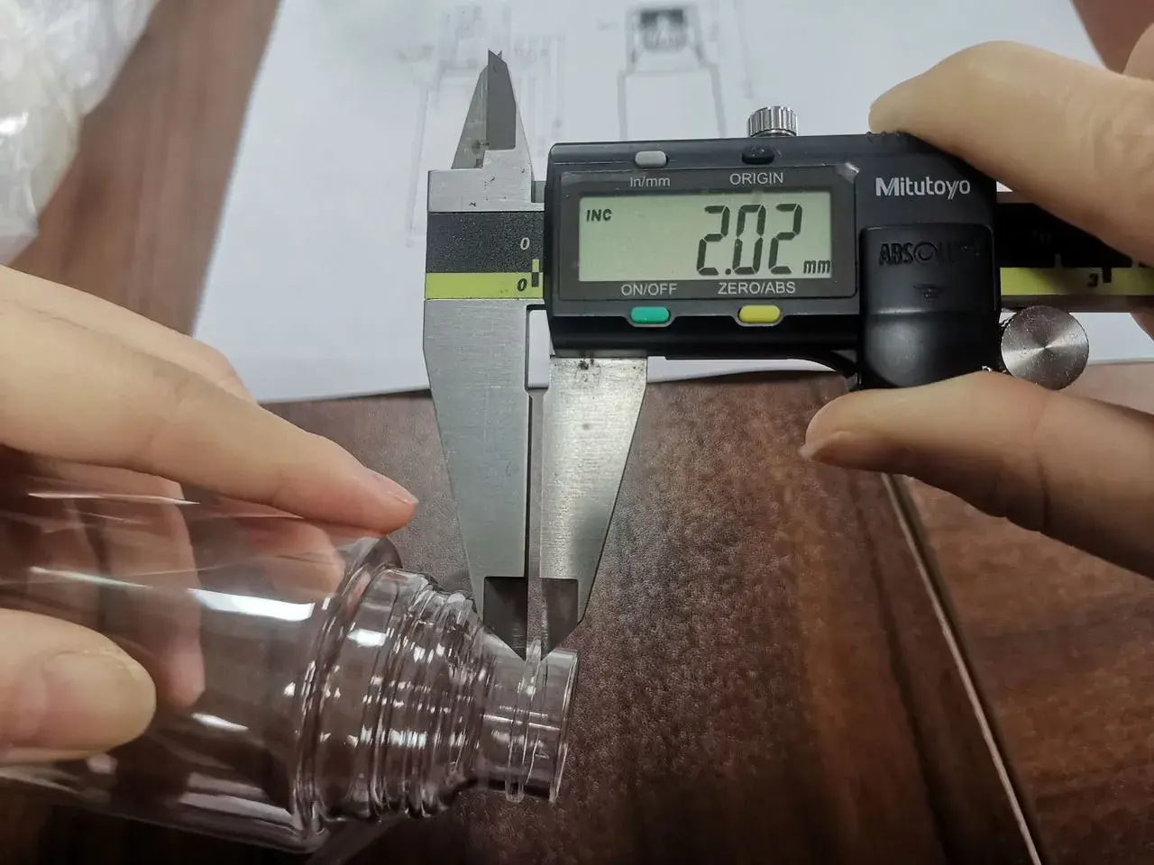 Digital caliper measuring the rim thickness of a clear plastic bottle during inspection.