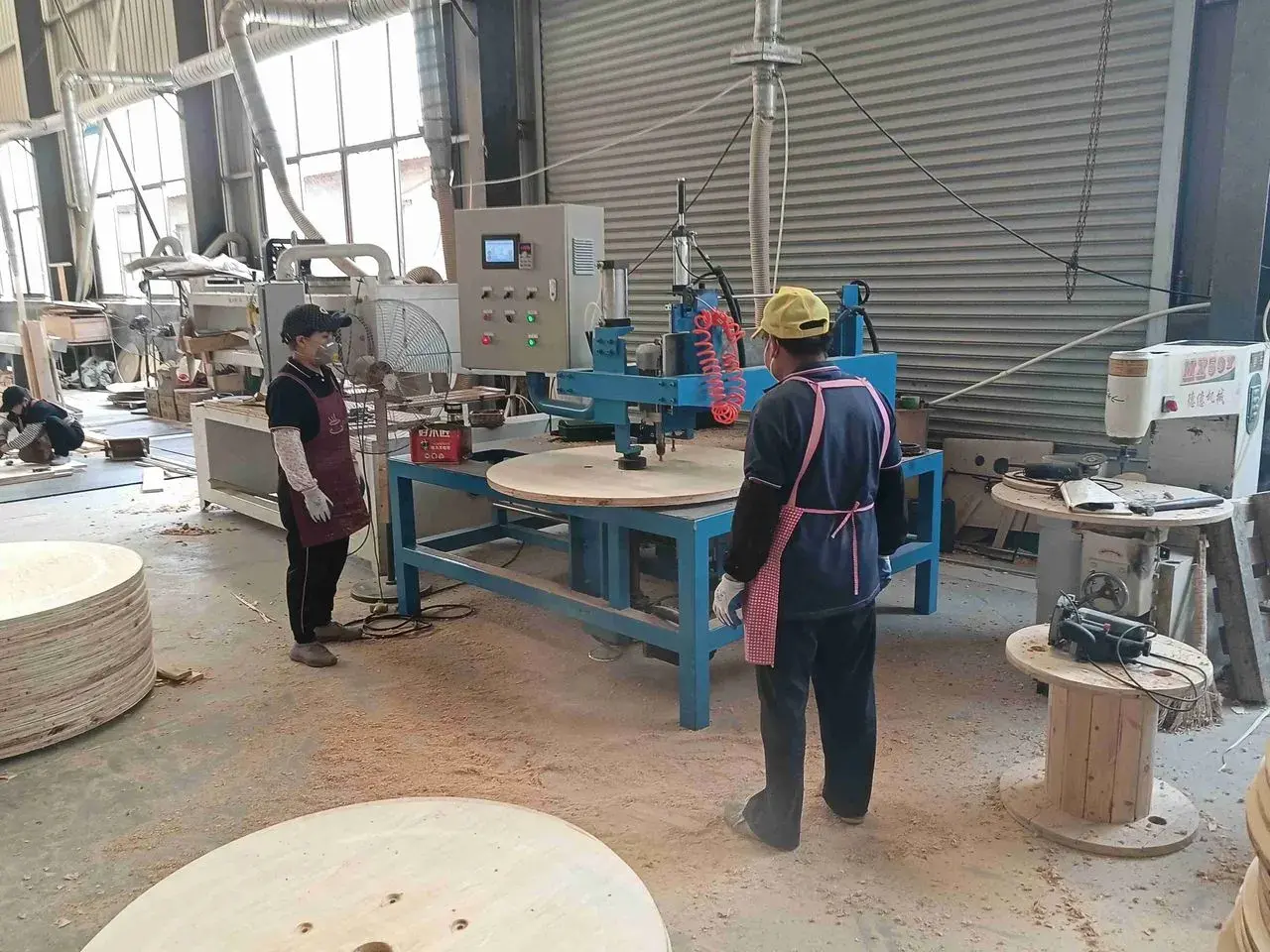Workers beside woodworking machinery and circular wooden panels in a factory workshop.